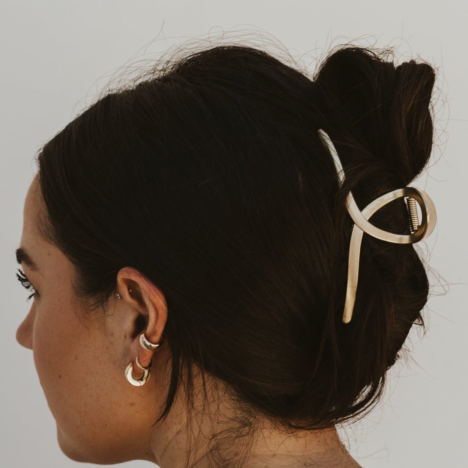 Girl with dark brown hair wearing a gold loop claw clip in her hair standing against a white background