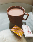 A brown ribbed ceramic mug sitting on top of a glass-top table holding hot chocolate. There are two retro matchboxes lying next to the mug