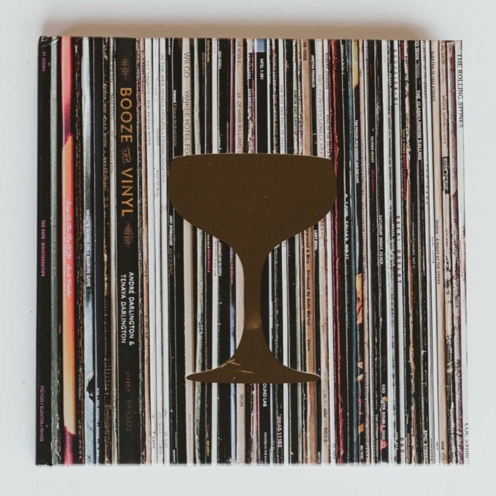 A square shaped book with photograph of vinyl stack for cover with gold coctail glass on top. Text on book reads, &quot;Booze and Vinyl. Photographed on white background. 