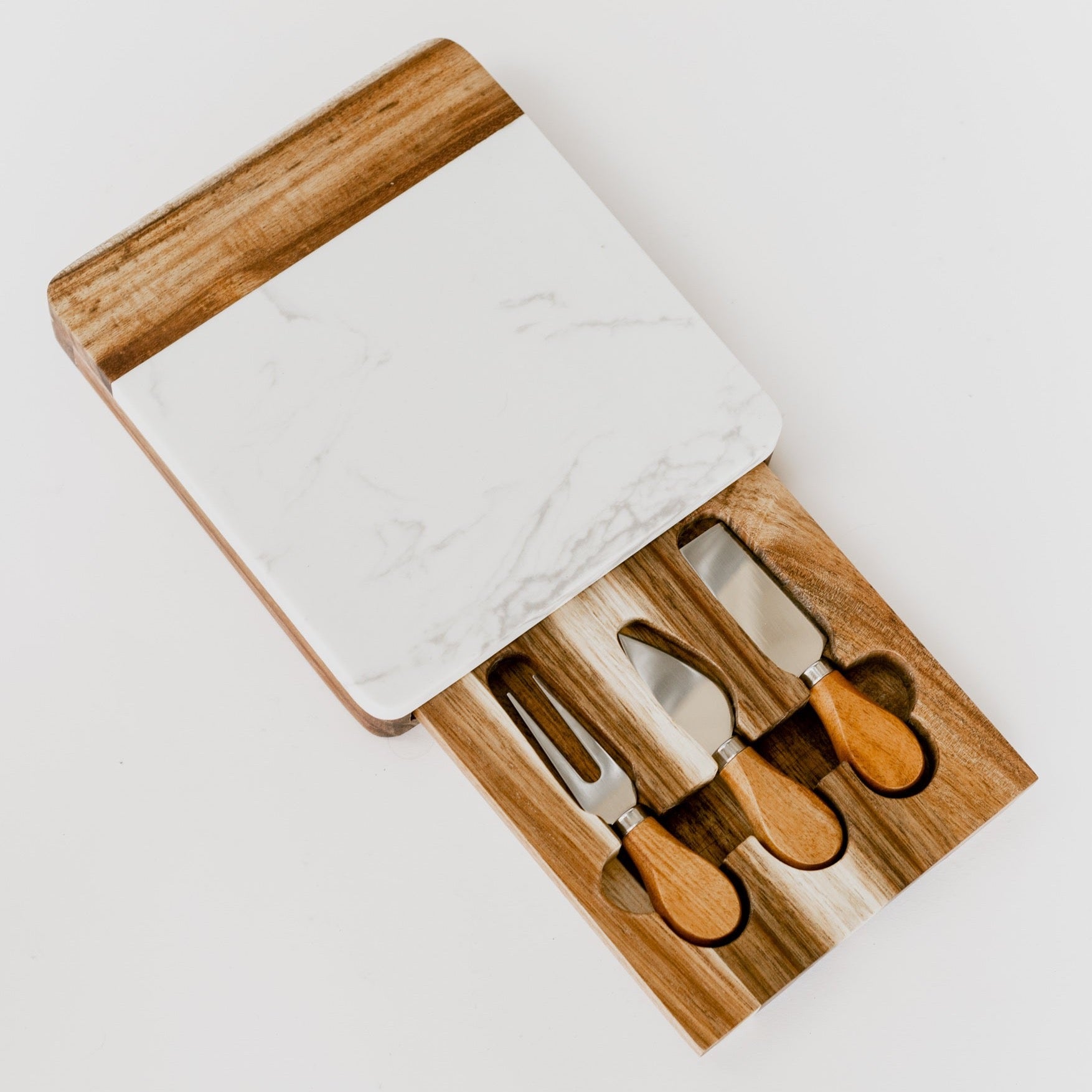 A white marble and acacia wood charcuterie board with a pull out drawer holding 3 stainless steel cheese knives against a white background