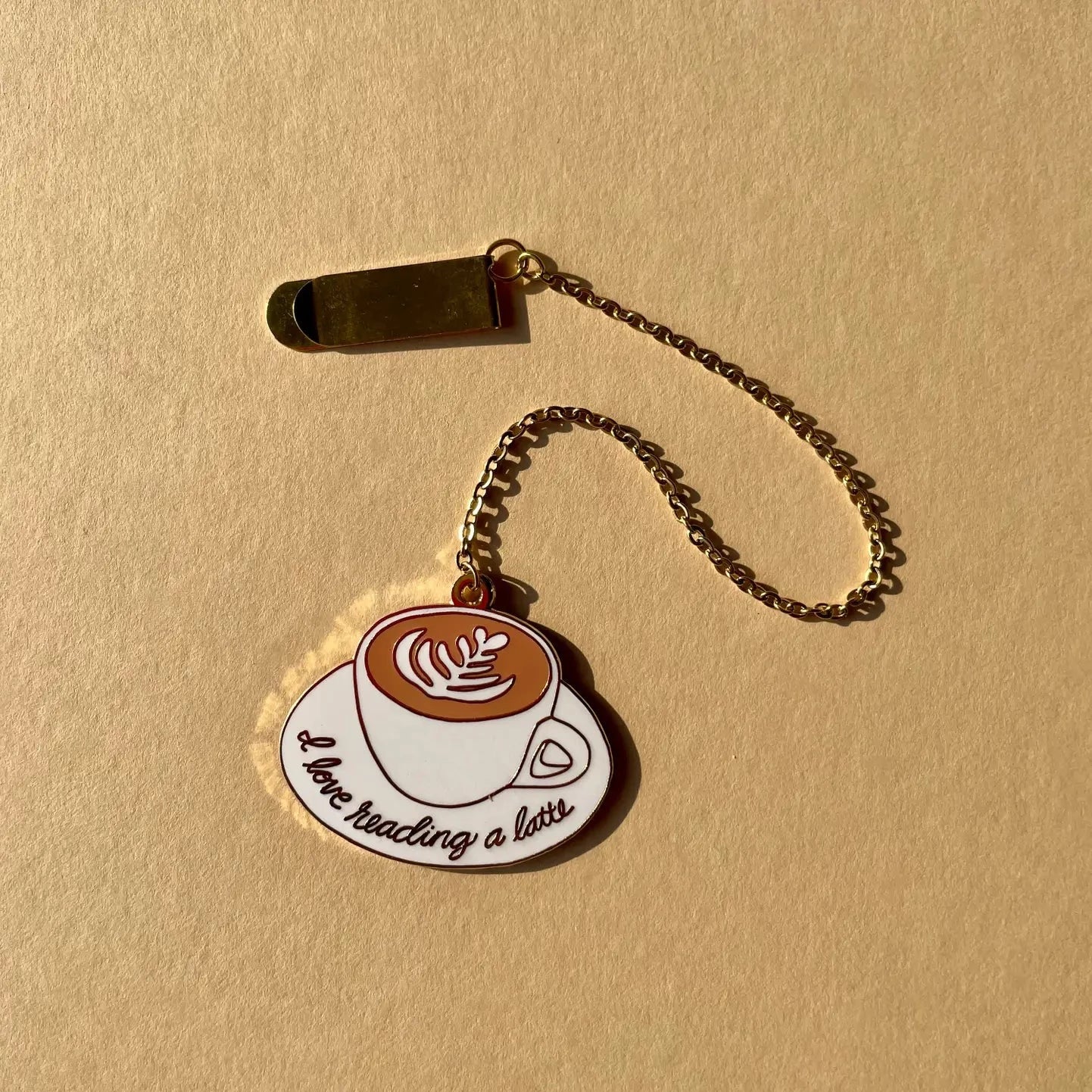 Enamel bookmark with gold chain and clip. Bookmark is a white cup with serving plate, inside the cup is a latter. Across the plate in gold text is printed "I Love reading a latte"