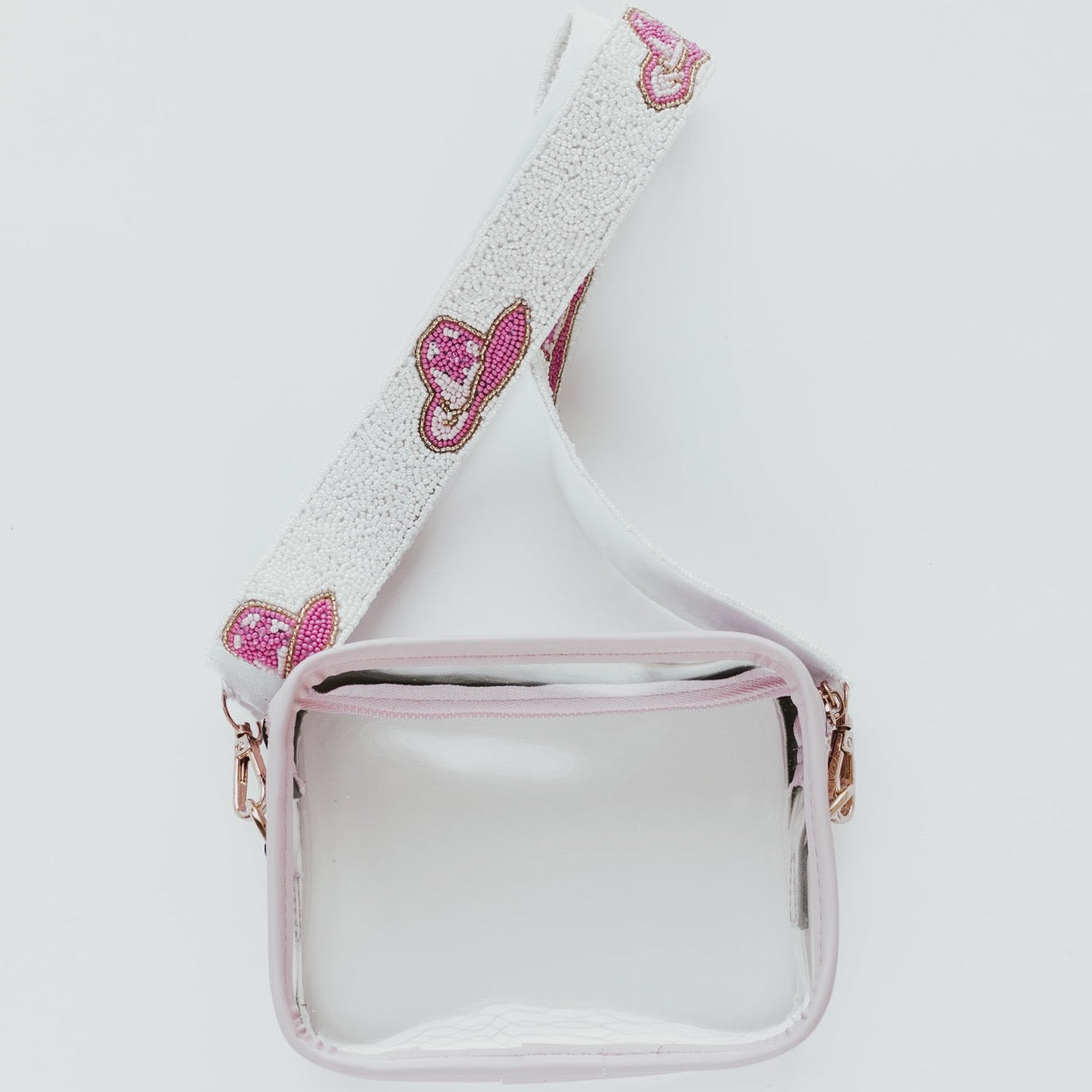 A clear purse with light pink lining and a white beaded bag strap with hot and light pink cowboy hats along the length of the strap against a white background
