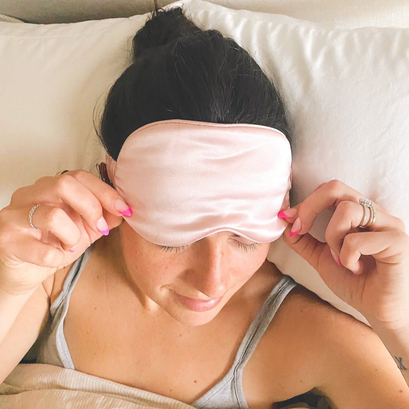 A girl laying in bed with her head on a white pillow and a pink silk sleep mask over her eyes.