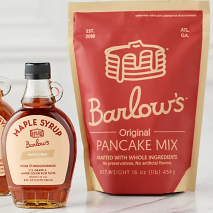 A bottle of maple syrup to the left of a red and beige plastic resealable bag of pancake mix photographed on white background.