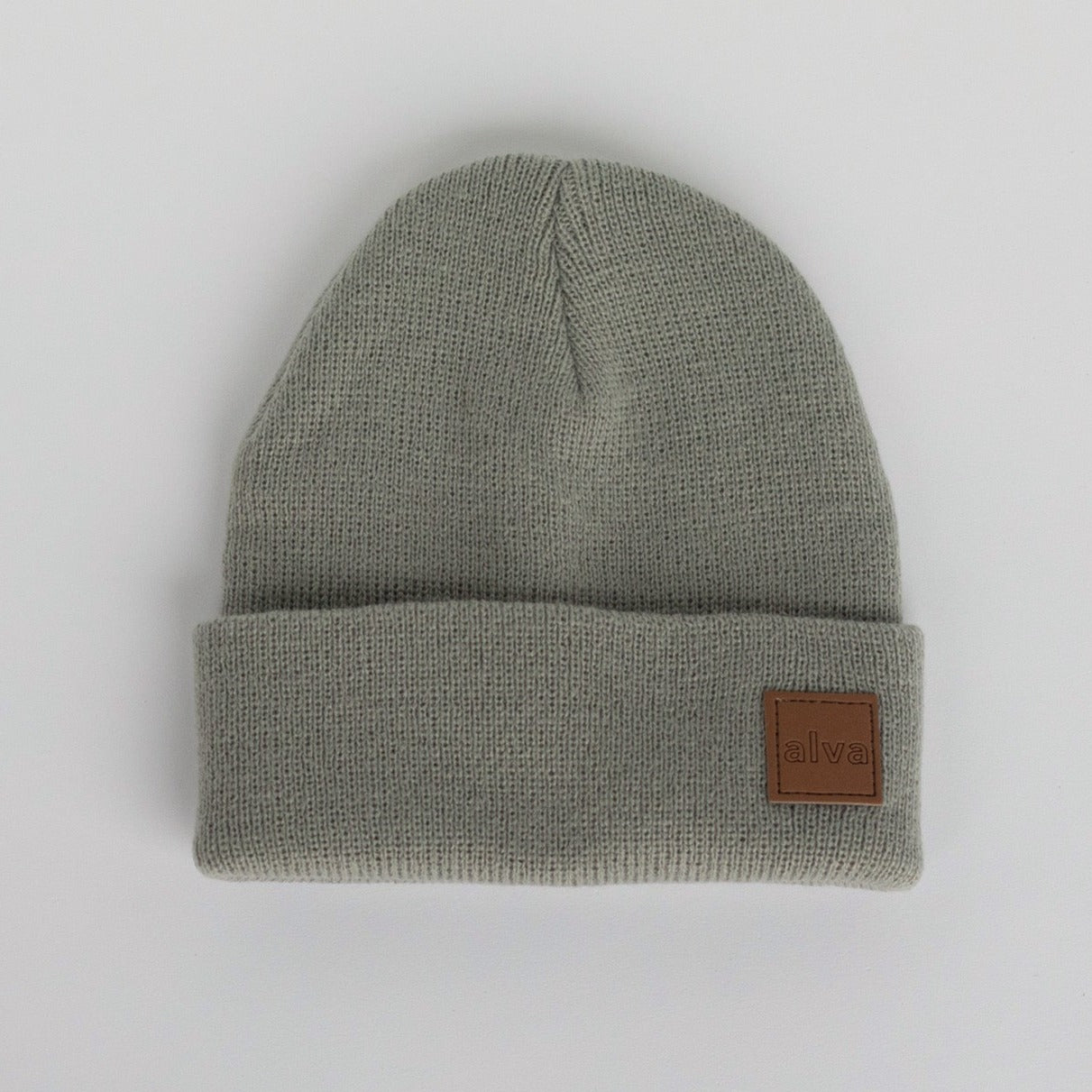 A sage grey colored ribbed baby beanie with a small square faux leather patch that says &quot;alva&quot; on the bottom right corner. The hat is sitting on a white background