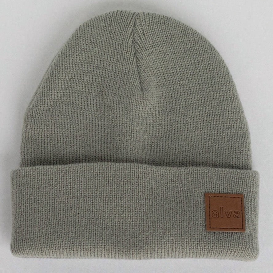 A sage grey colored ribbed baby beanie with a small square faux leather patch that says "alva" on the bottom right corner. The hat is sitting on a white background